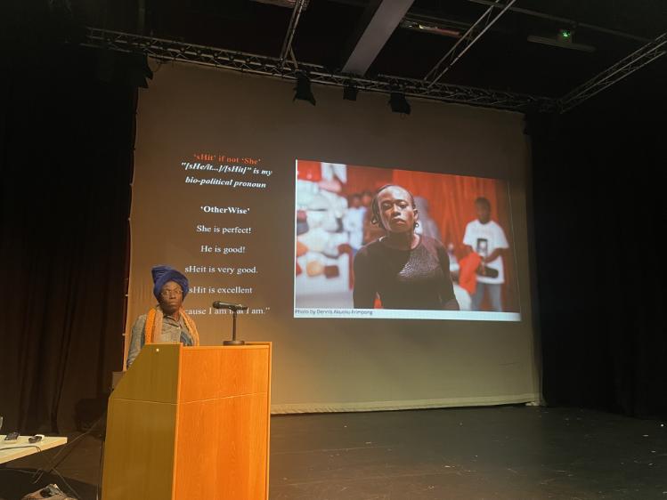 Va-Bene standing at a lectern in the Brian Friel in front of large screen displaying presentation